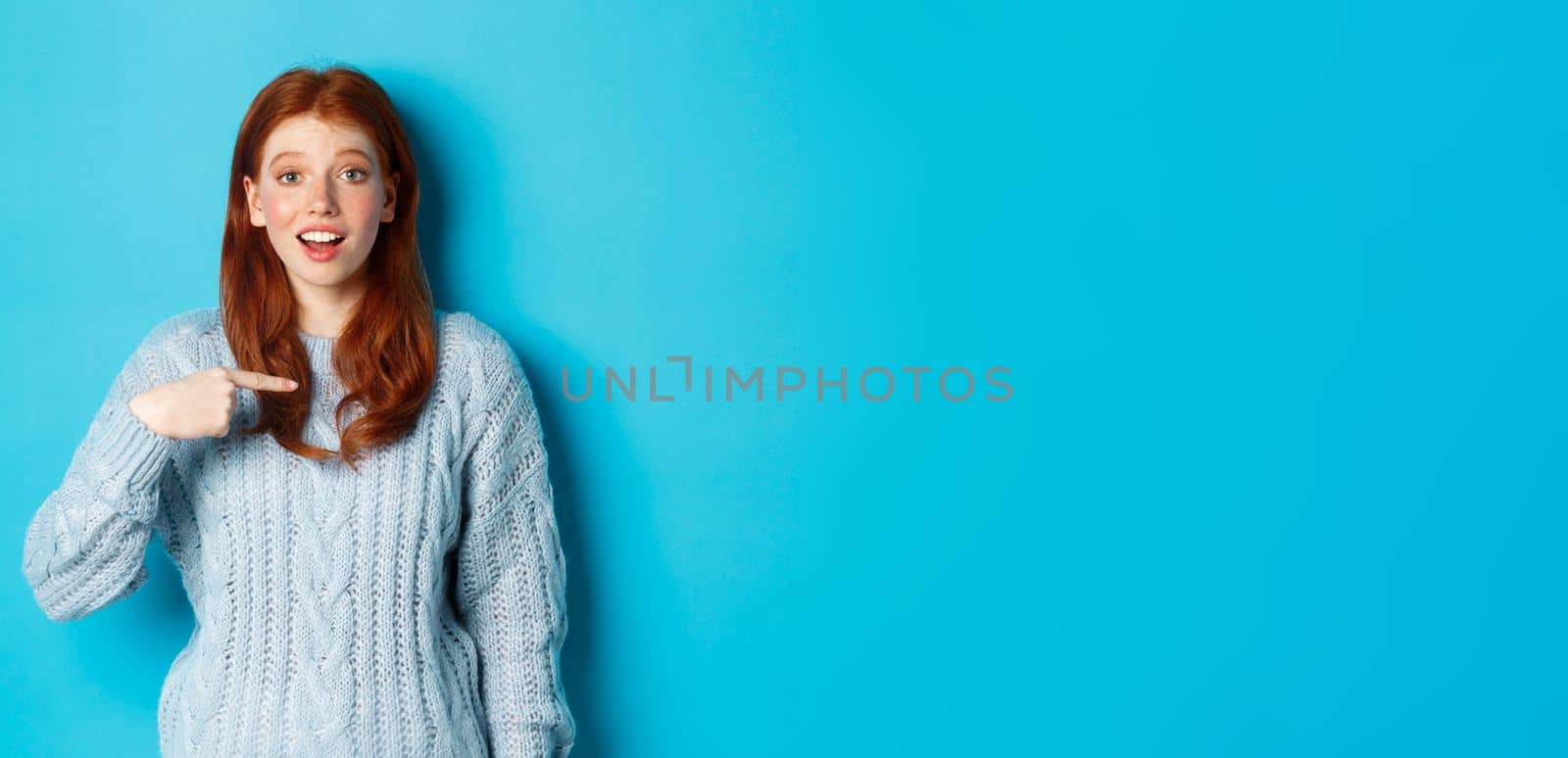 Hopeful redhead girl pointing at herself, standing over blue background.