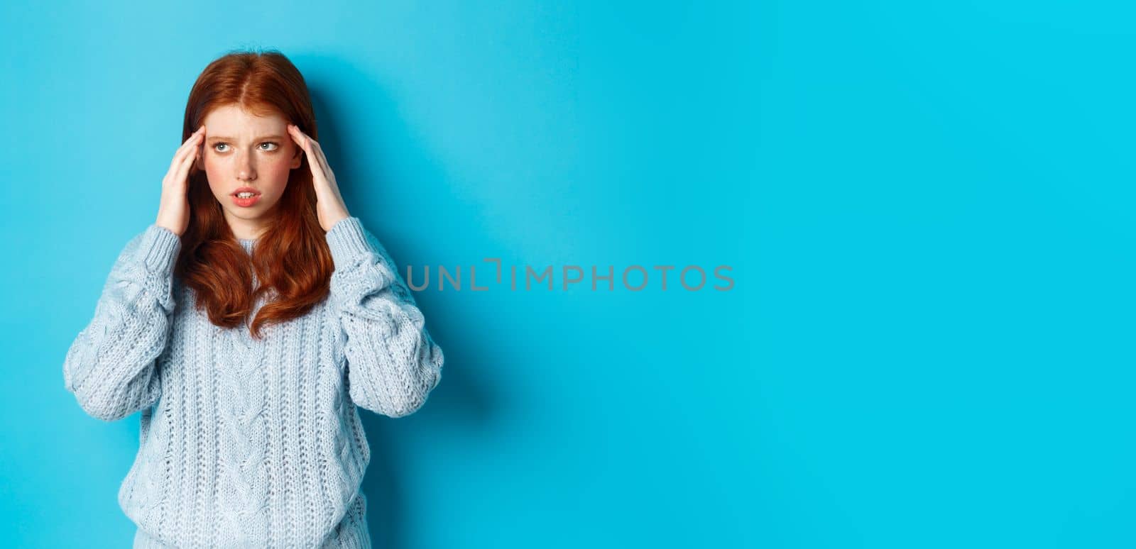 Young tensed redhead college girl trying to think, rubbing temples and looking up, thinking, standing over blue background.