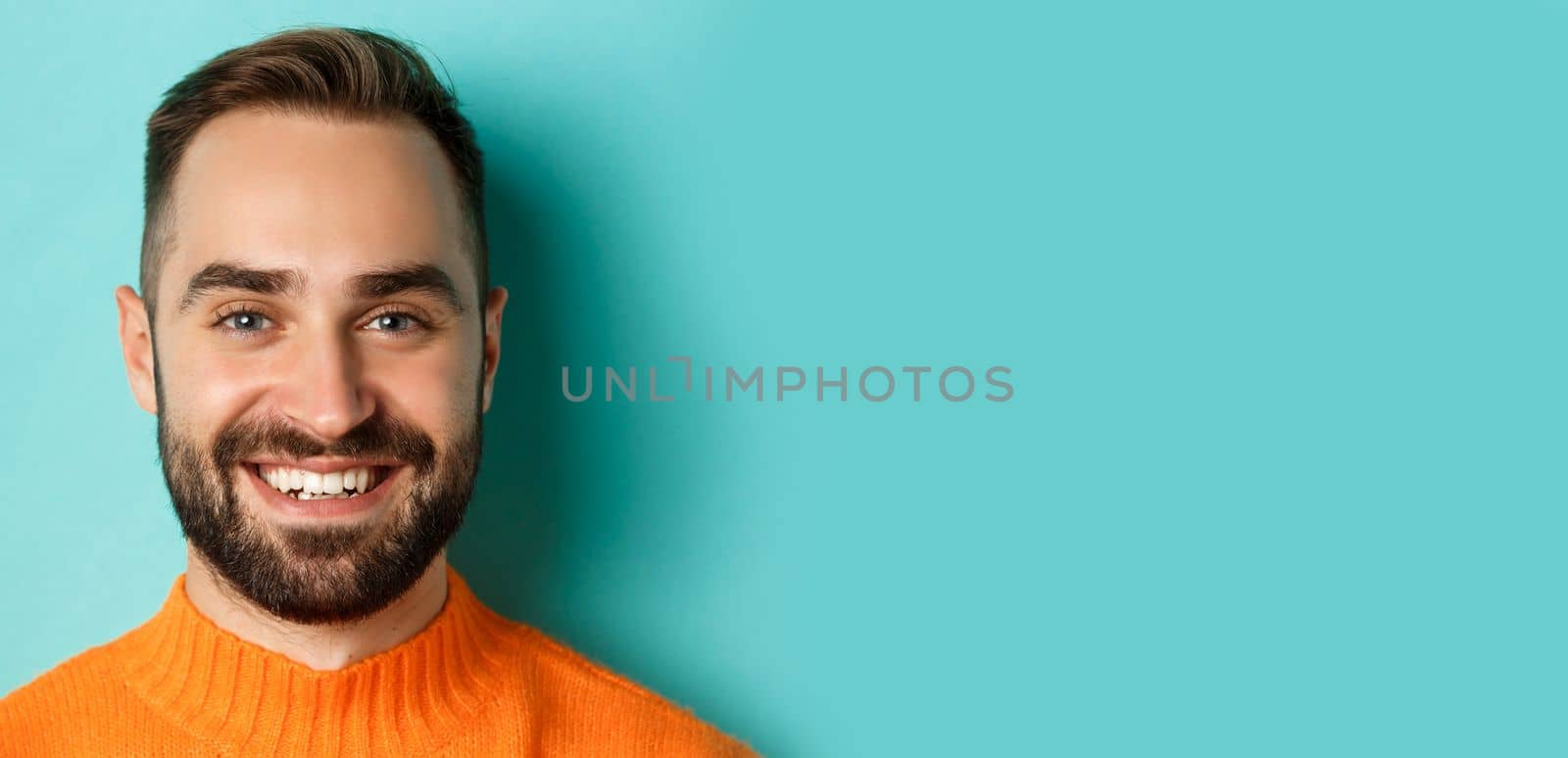 Headshot of handsome caucasian man with beard smiling happy at camera, standing in orange sweater against turquoise background.