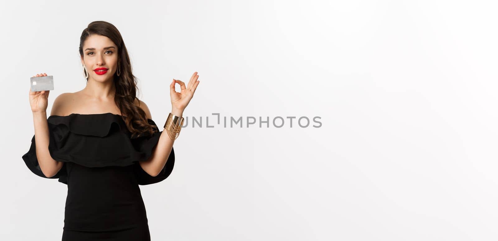 Beauty and shopping concept. Gorgeous woman in luxury jewelry and black dress, showing okay sign and credit card, standing over white background.