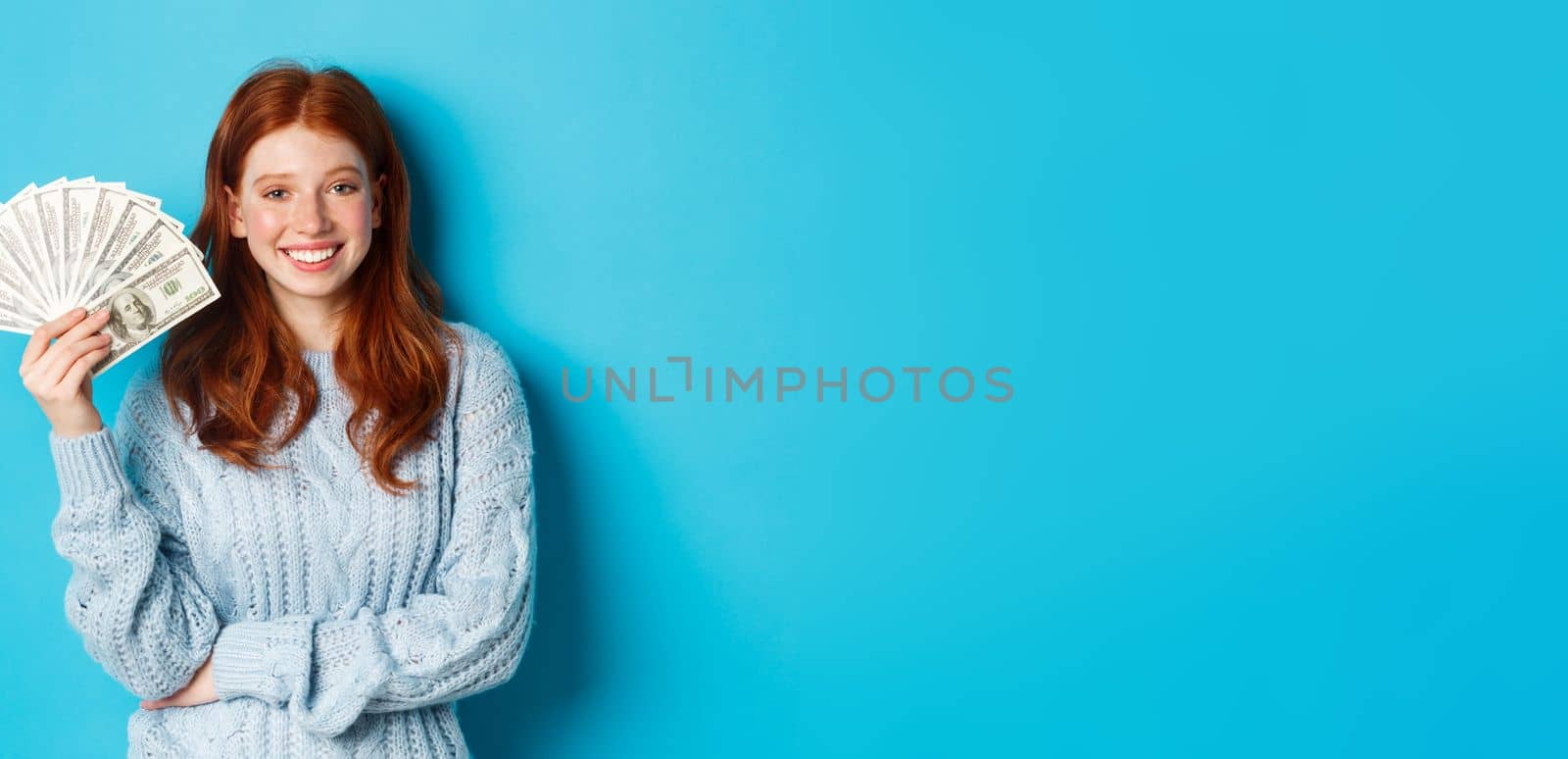 Happy and unbothered redhead woman holding money and smiling, looking confident and carefree, standing in sweater against blue background by Benzoix