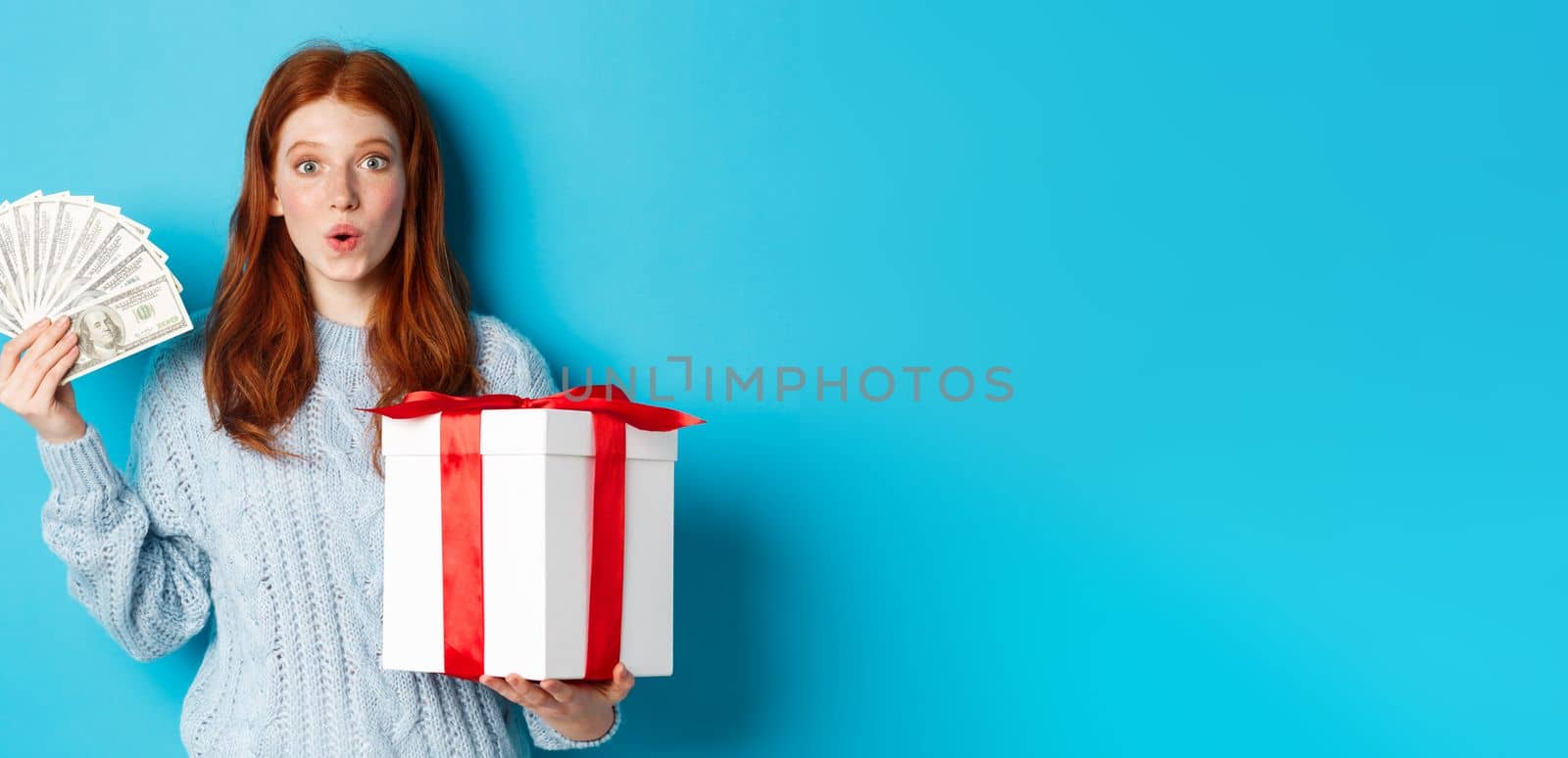 Christmas and shopping concept. Excited redhead girl looking at camera, holding big New Year gift and dollars, buying presents, standing over blue background.