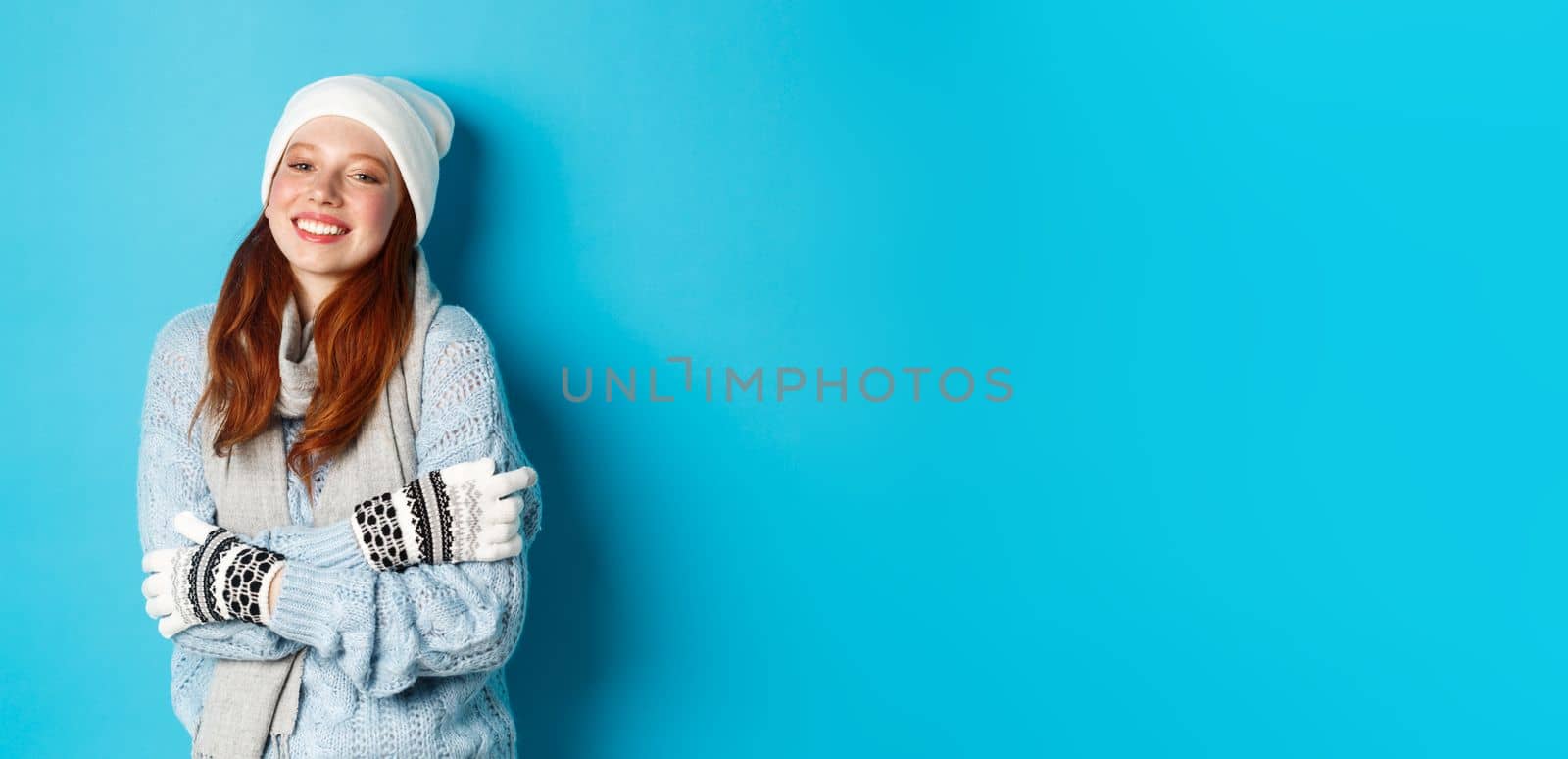 Winter and holidays concept. Smiling redhead girl in beanie, gloves and sweater getting warm after going outside, standing over blue background by Benzoix