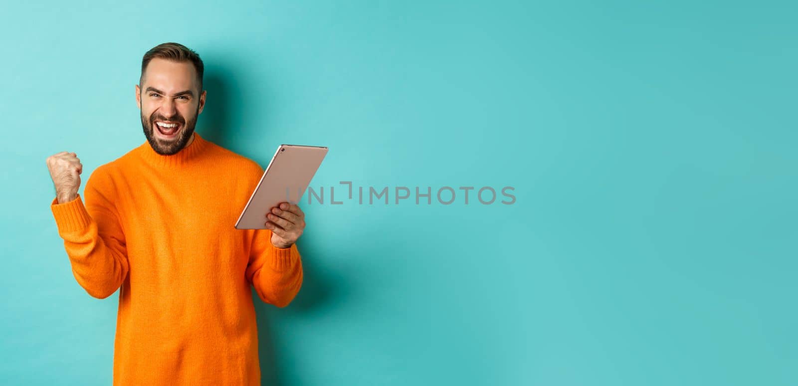 Cheerful winning man holding digital tablet, rejoicing and celebrating victory in game, making fist pump gesture, standing over light blue background by Benzoix