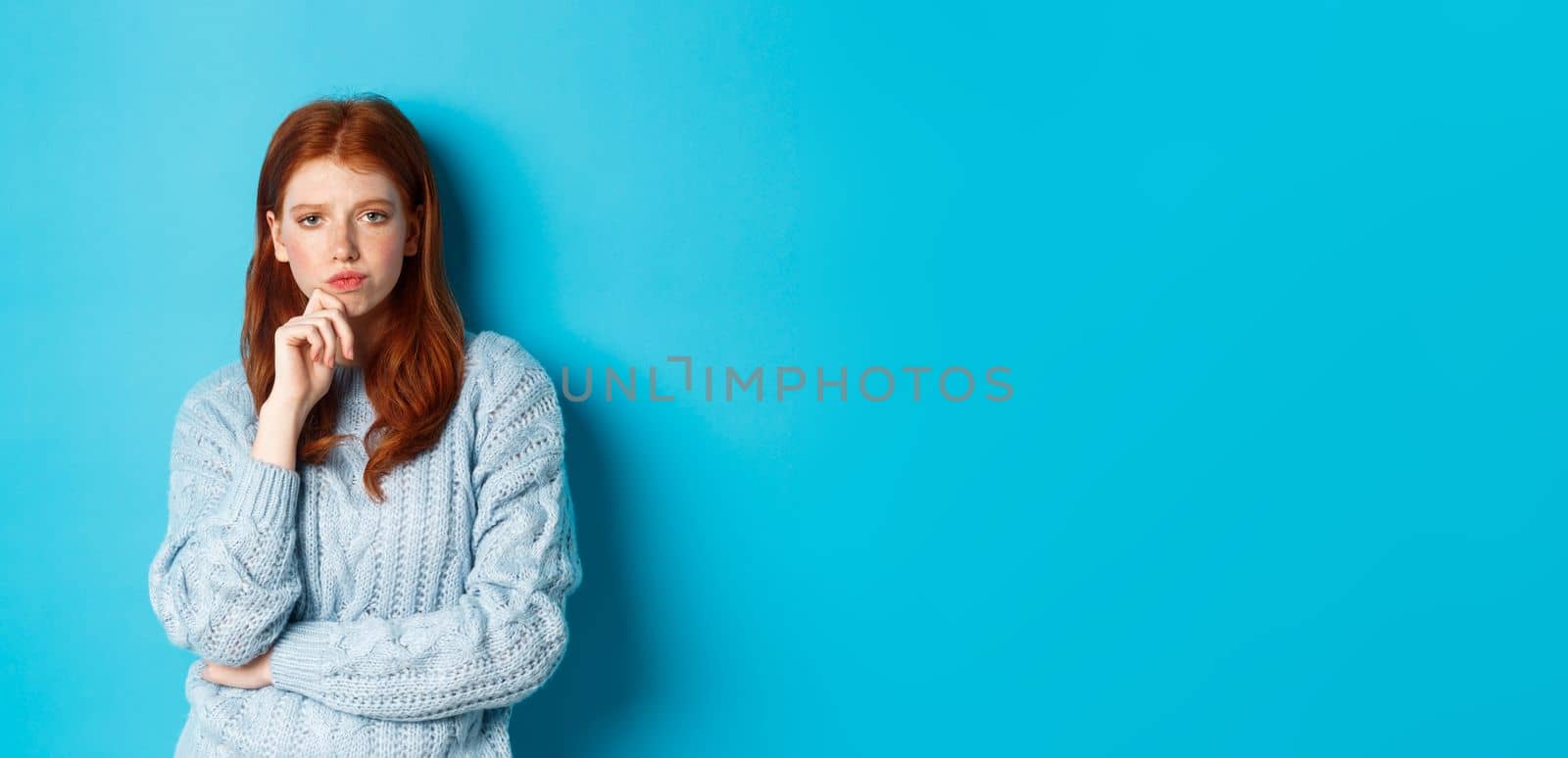 Thoughtful teenage redhead girl staring at camera, pondering ideas, making decision with serious face, standing over blue background by Benzoix