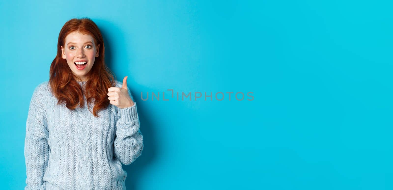 Impressed redhead girl showing thumb up and staring amazed at camera, praising good work, standing amazed against blue background by Benzoix