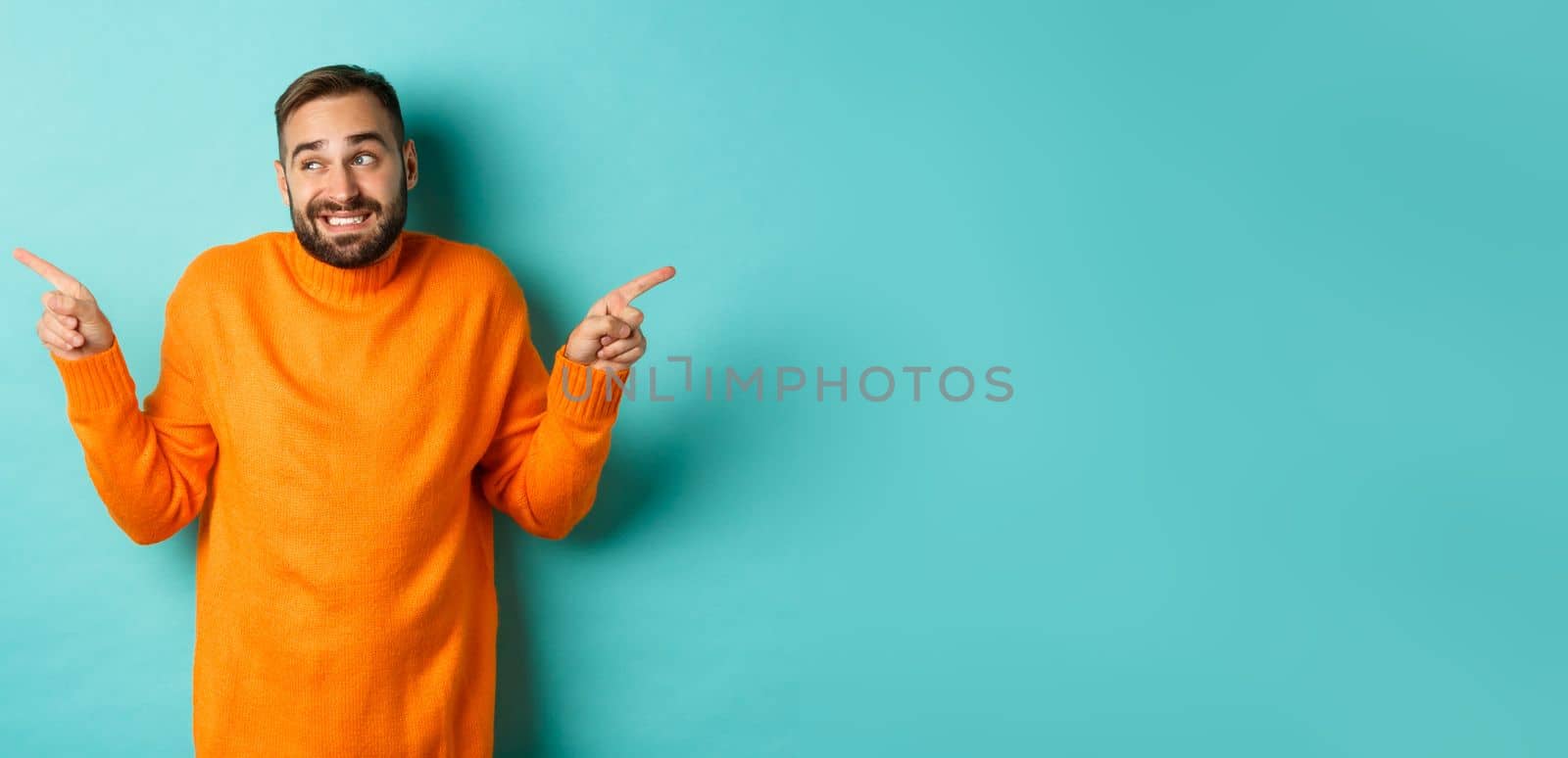 Confused handsome man shrugging indecisive, pointing fingers sideways, cant choose between two variants, standing against light blue background.