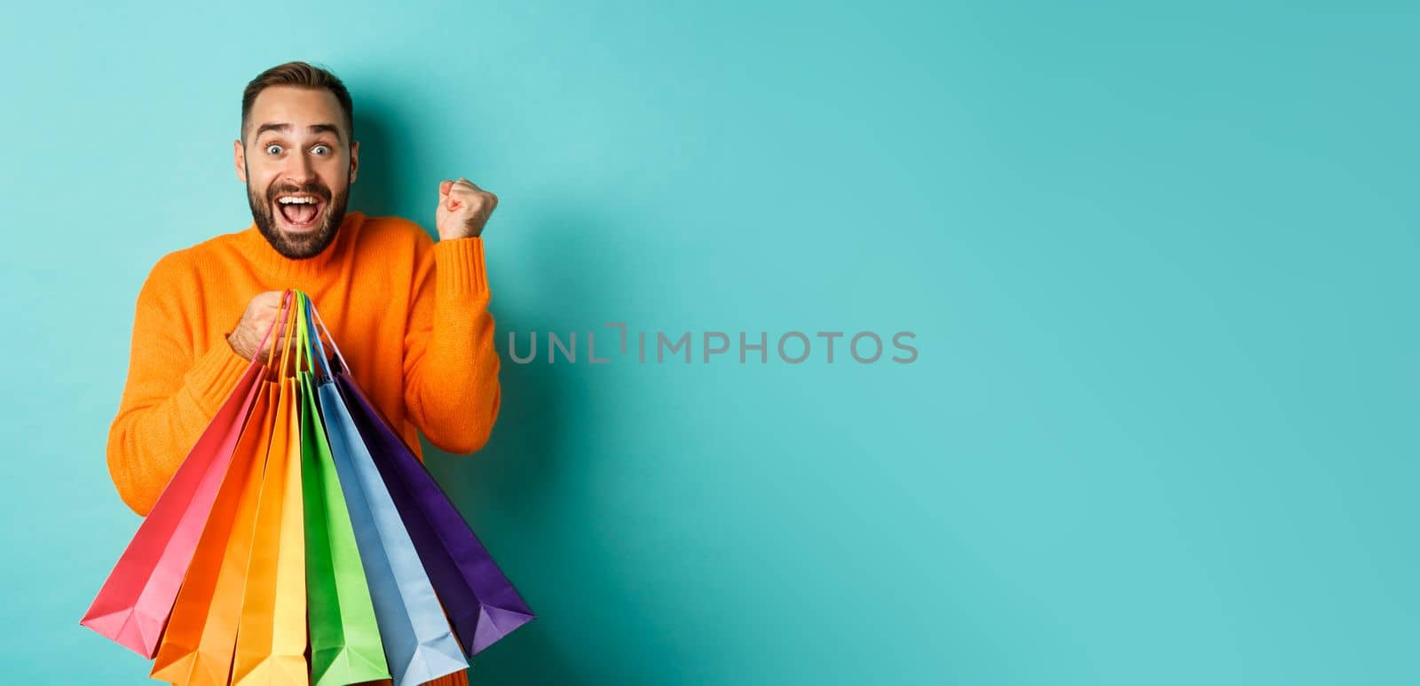 Lifestyle concept. Excited man showing shopping bags and rejoicing from discounts, standing over turquoise background by Benzoix