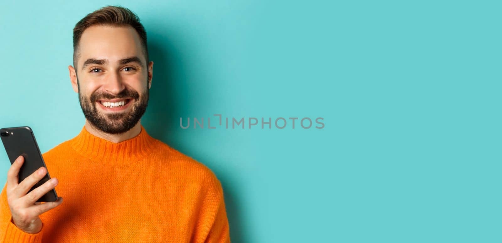 Close-up of happy handsome man writing message on mobile phone, holding smartphone and smiling, standing against turquoise background by Benzoix