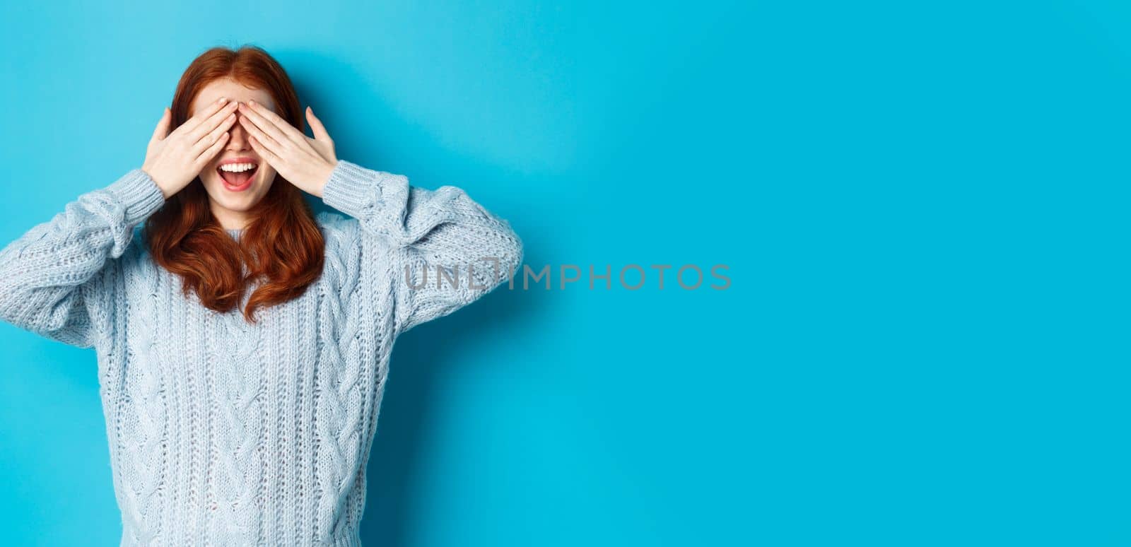 Cheerful redhead female model close eyes and waiting for christmas gift, holding hands on face and smiling amused, anticipating surprise, standing over blue background.