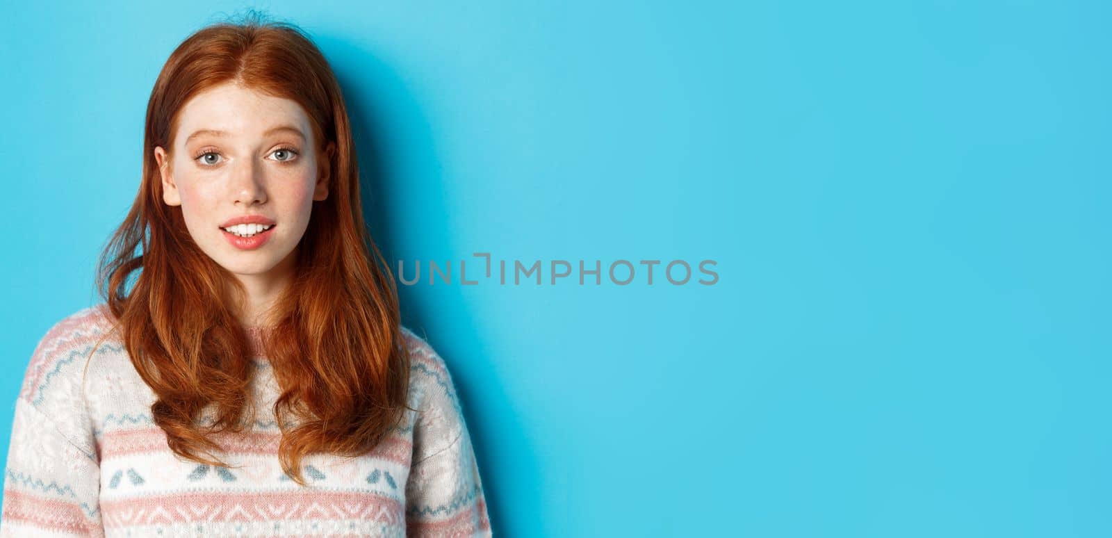 Close-up of redhead girl looking intrigued at camera, hear something interesting, standing over blue background.