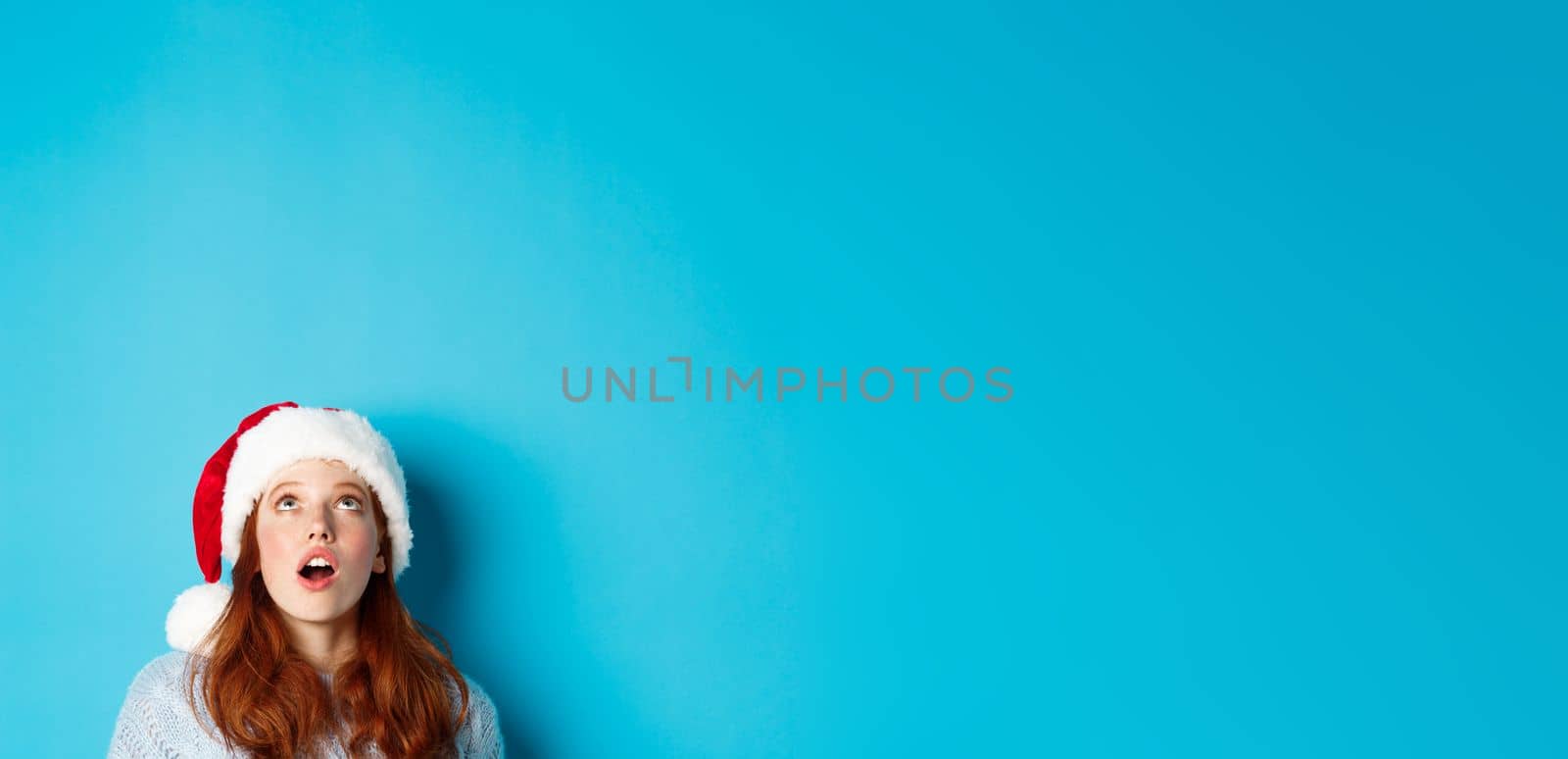 Winter holidays and Christmas eve concept. Head of pretty redhead girl in santa hat, appear from bottom and looking up at logo, seeing promo offer amazed, blue background by Benzoix