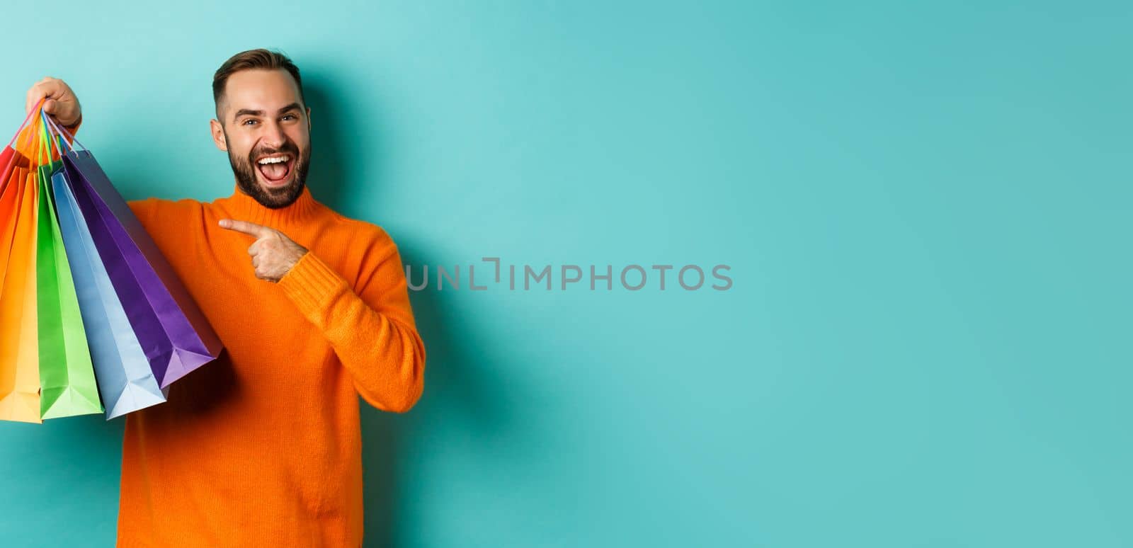 Happy handsome man holding shopping bags and smiling, pointing at purchased items and recommending store, standing over turquoise background.