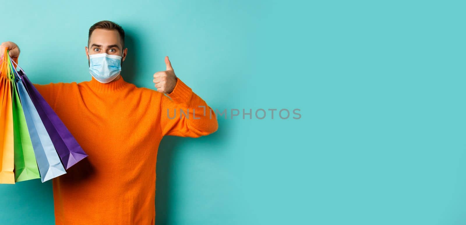 Covid-19, social distancing and lifestyle concept. Young man in face mask showing shopping bags and thumb-up, buying holiday gifts during pandemic by Benzoix