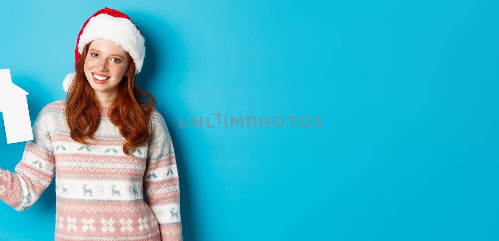 Holiday promos and real estate concept. Cheerful redhead woman in santa hat holding paper house in hand and smiling, standing in sweater against blue background by Benzoix