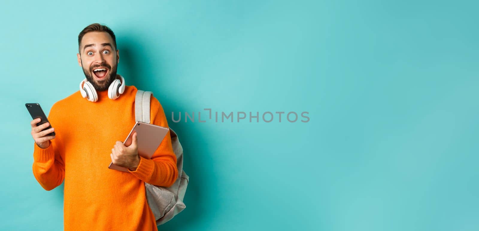 Handsome man student with headphones and backpack, holding digital tablet and smartphone, looking amazed at camera, standing against turquoise background by Benzoix