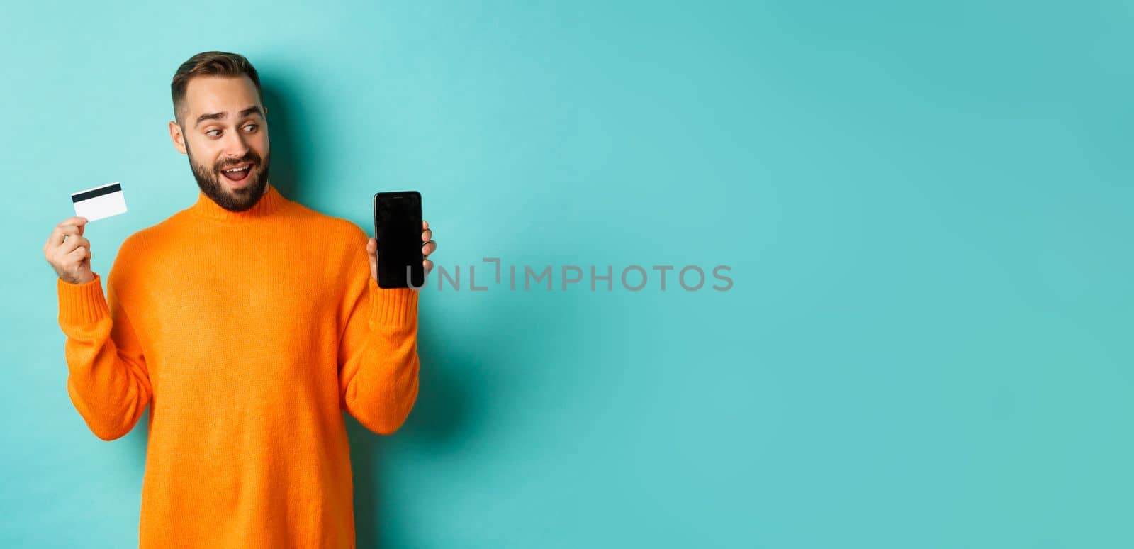 Online shopping. Amazed guy using credit card and showing mobile screen, looking impressed, standing against light blue background.