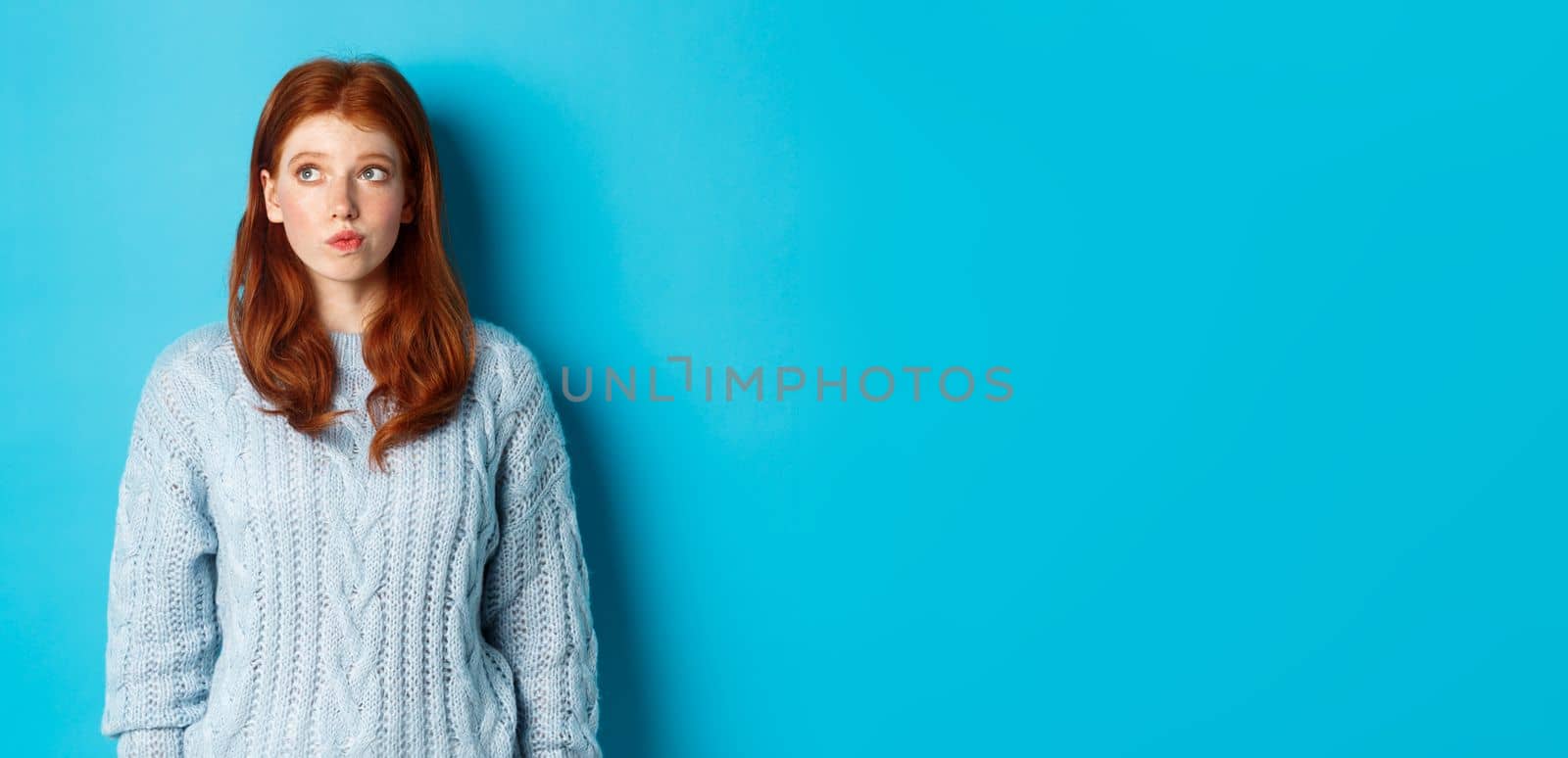 Dreamy redhead girl thinking or making decision, looking at upper left corner logo, standing against blue background by Benzoix