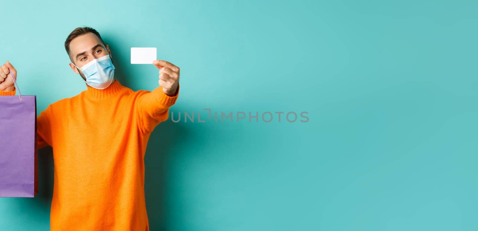 Covid-19, pandemic and lifestyle concept. Happy male customer in face mask showing credit card and purple shopping bag, standing over light blue background by Benzoix