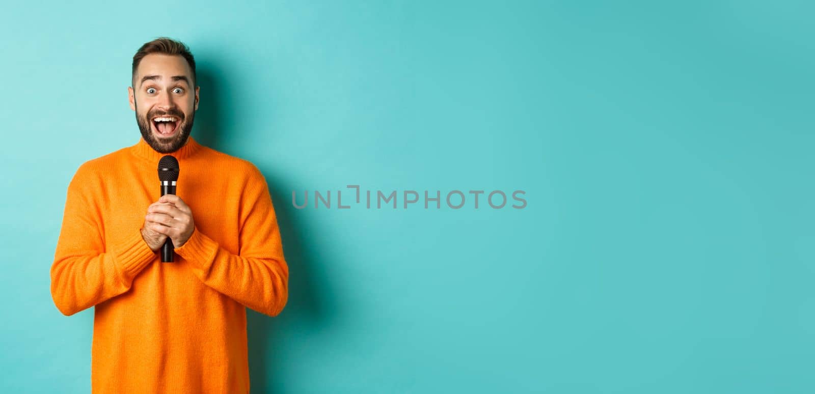 Happy adult man singing karaoke, holding microphone and looking at camera, standing in orange sweater against turquoise background by Benzoix