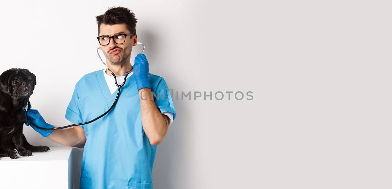 Confused male doctor veterinarian checking dog with stethoscope, looking puzzled, standing over white background.