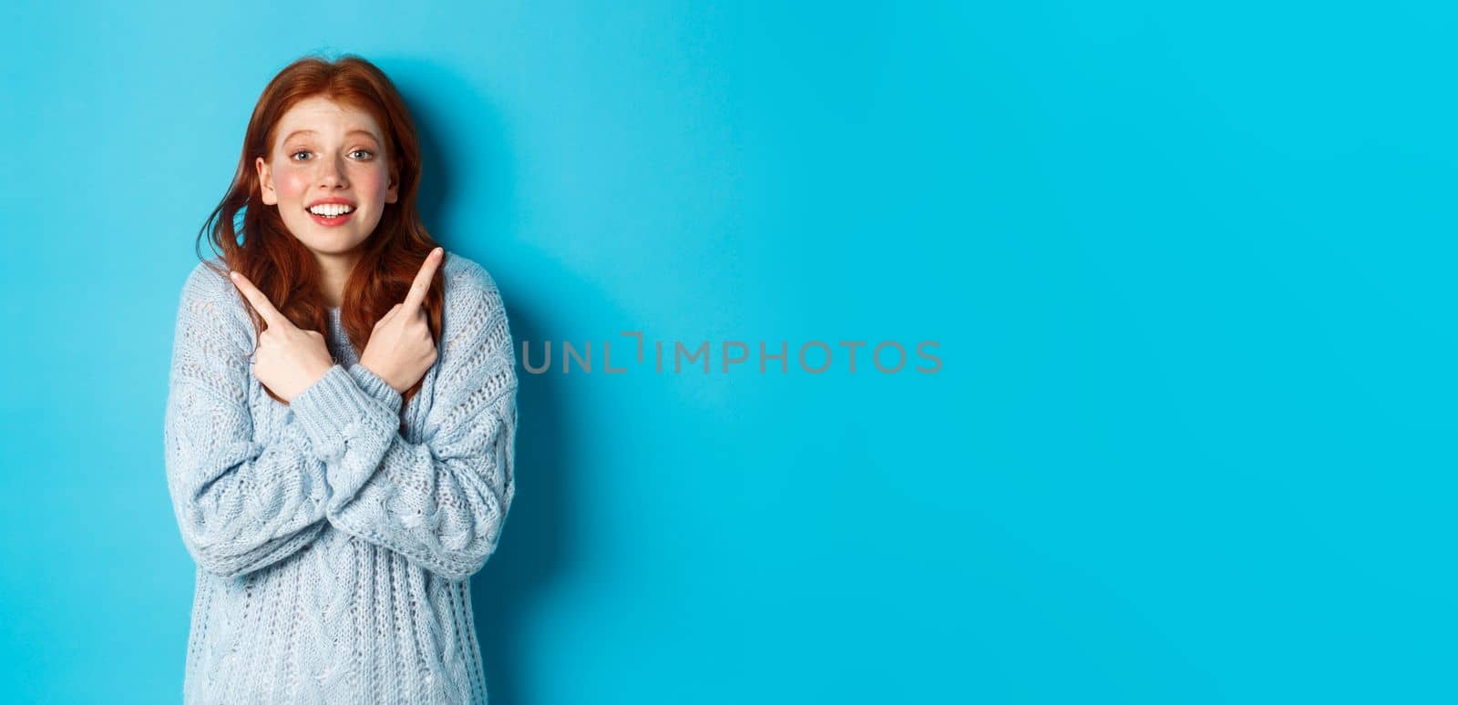 Excited redhead girl pointing fingers sideways, showing two choices and looking tempted at camera, standing against blue background.