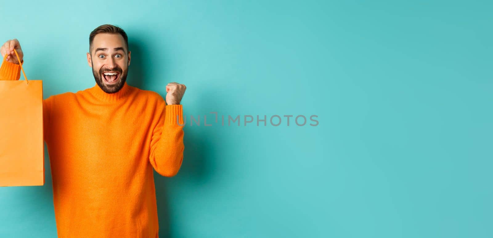 Happy man holding orange shopping bag and rejoicing, got discount and celebrating, standing over turquoise background.