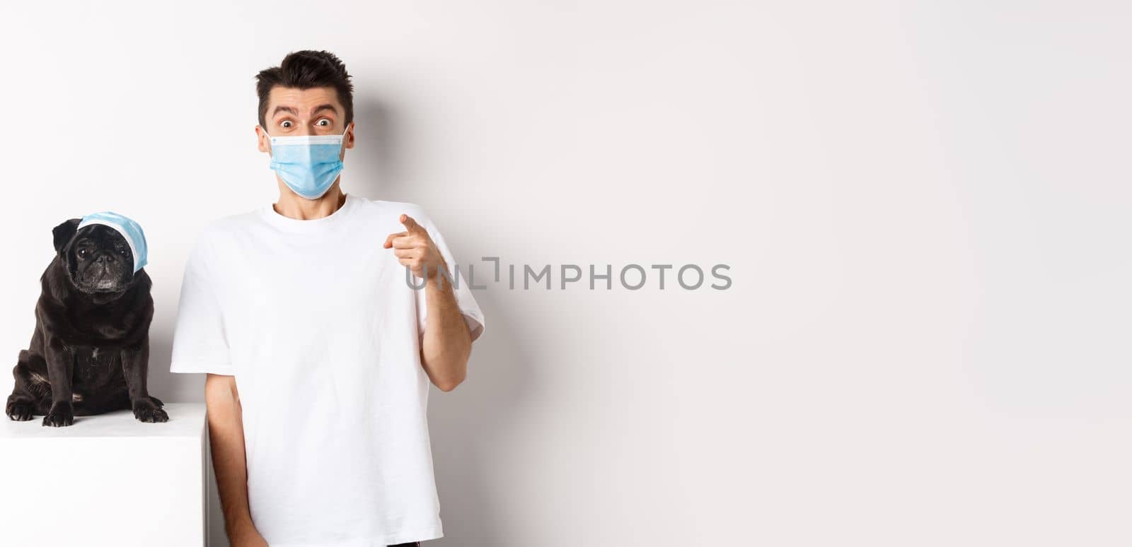 Covid-19, animals and quarantine concept. Happy dog owner and cute pug wearing medical masks, man pointing finger at camera amazed, white background by Benzoix