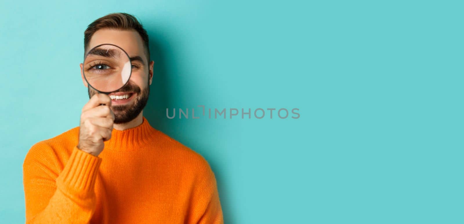 Funny man looking through magnifying glass, searching or investigating something, standing in orange sweater against turquoise background.