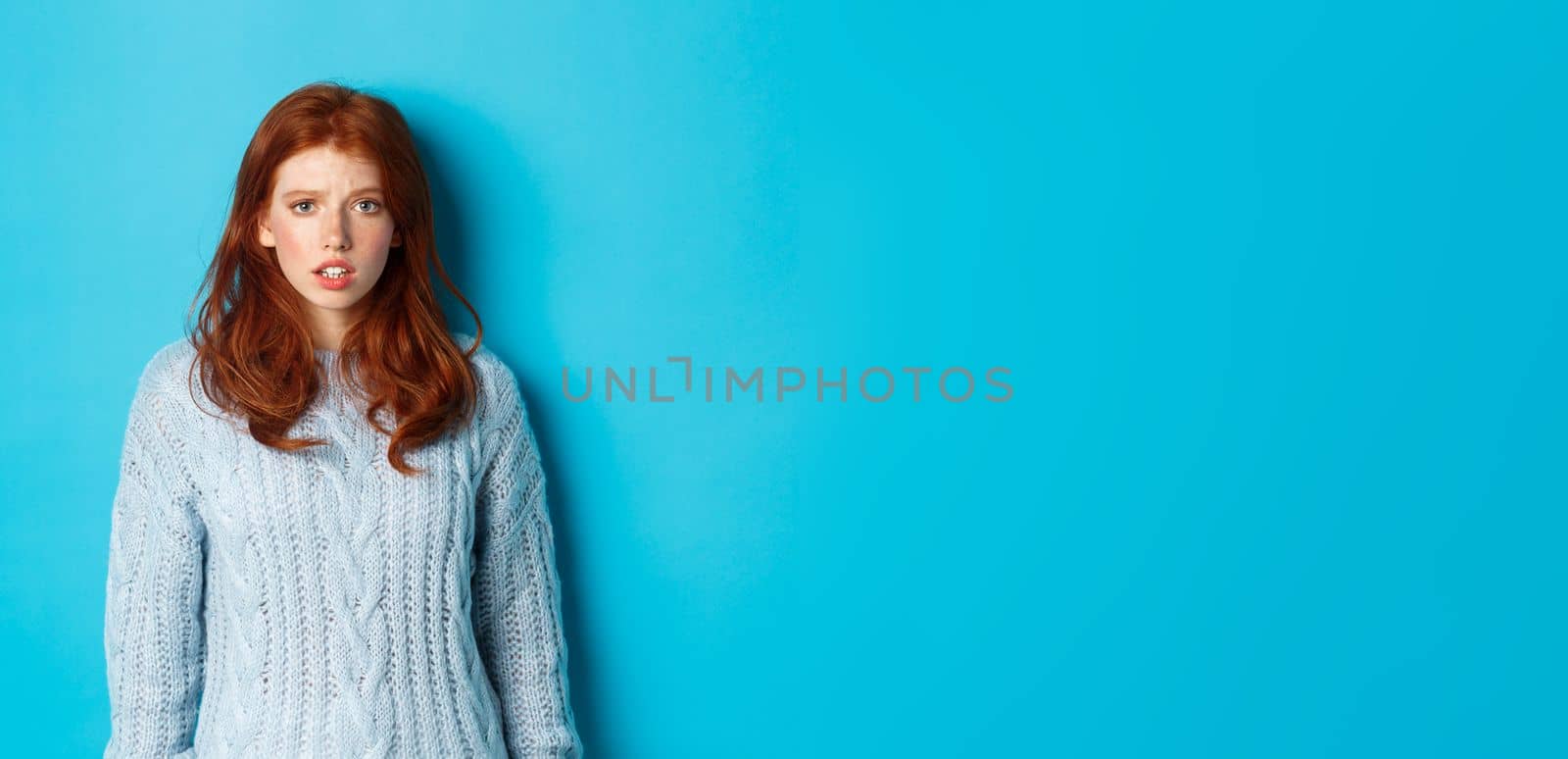 Confused redhead girl in sweater staring at camera, raising eyebrow and feeling puzzled, standing over blue background by Benzoix