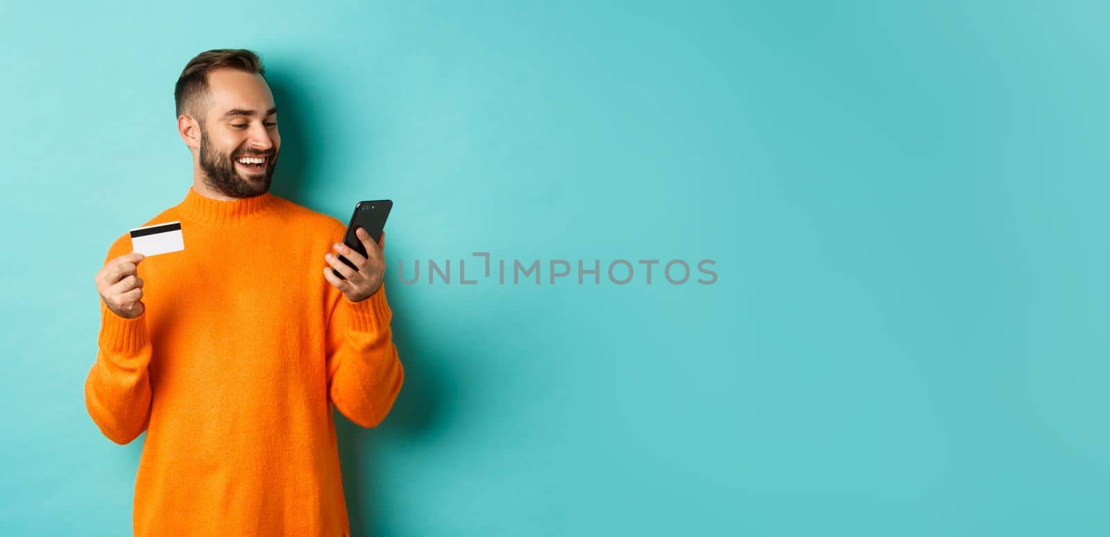 Online shopping. Handsome bearded man paying in internet, holding credit card and stare at mobile screen, standing over turquoise background.