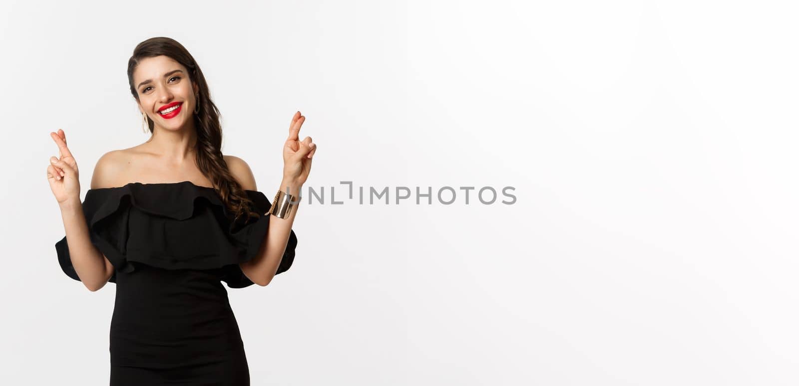 Fashion and beauty. Stylish glamour woman in black dress, red lips, looking optimistic and smiling while cross fingers, making wish, standing over white background.