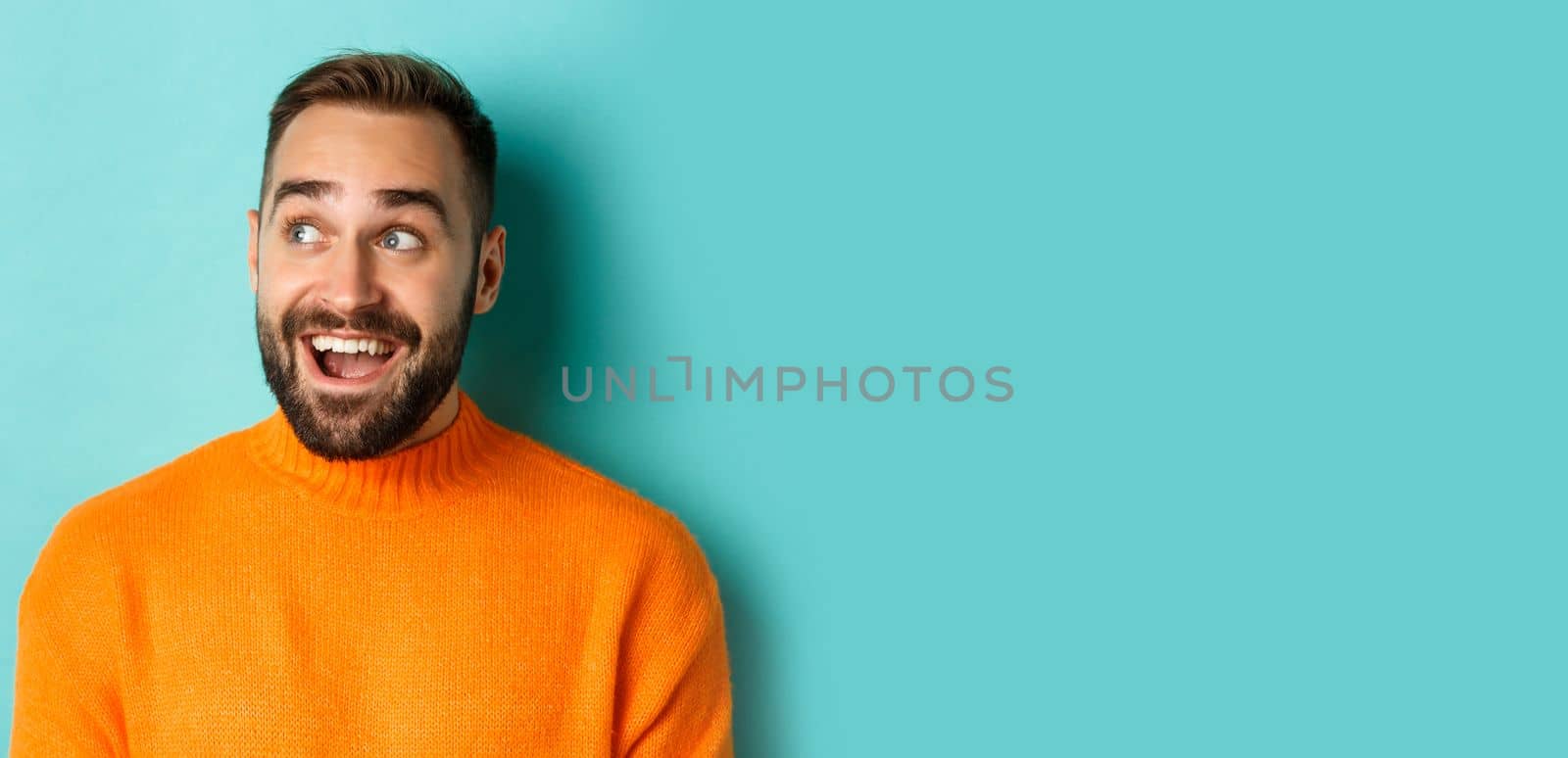 Close-up of handsome caucasian man smiling, looking left with surprised face, staring at logo, wearing orange sweater, standing against turquoise background.