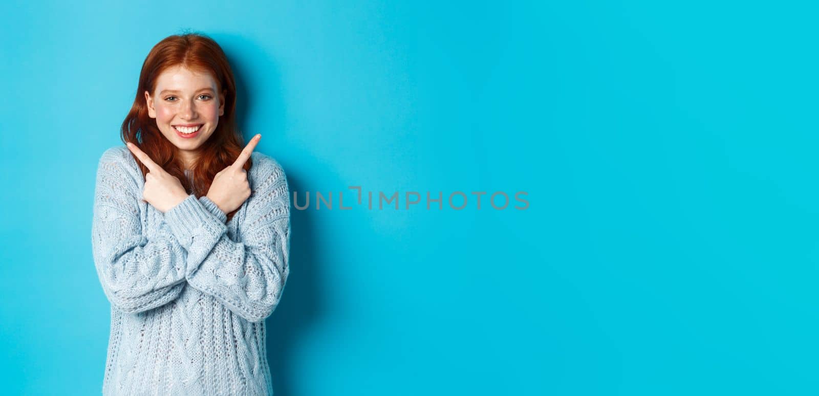 Cheerful redhead girl showing two choices, pointing sideways and smiling over blue background. Copy space