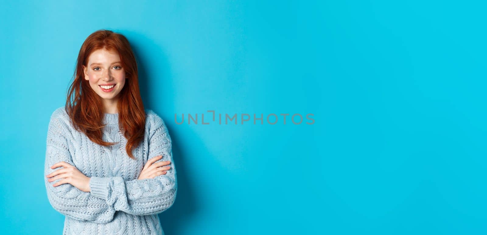 Attractive redhead girl in sweater smiling and staring at camera, standing confident against blue background by Benzoix