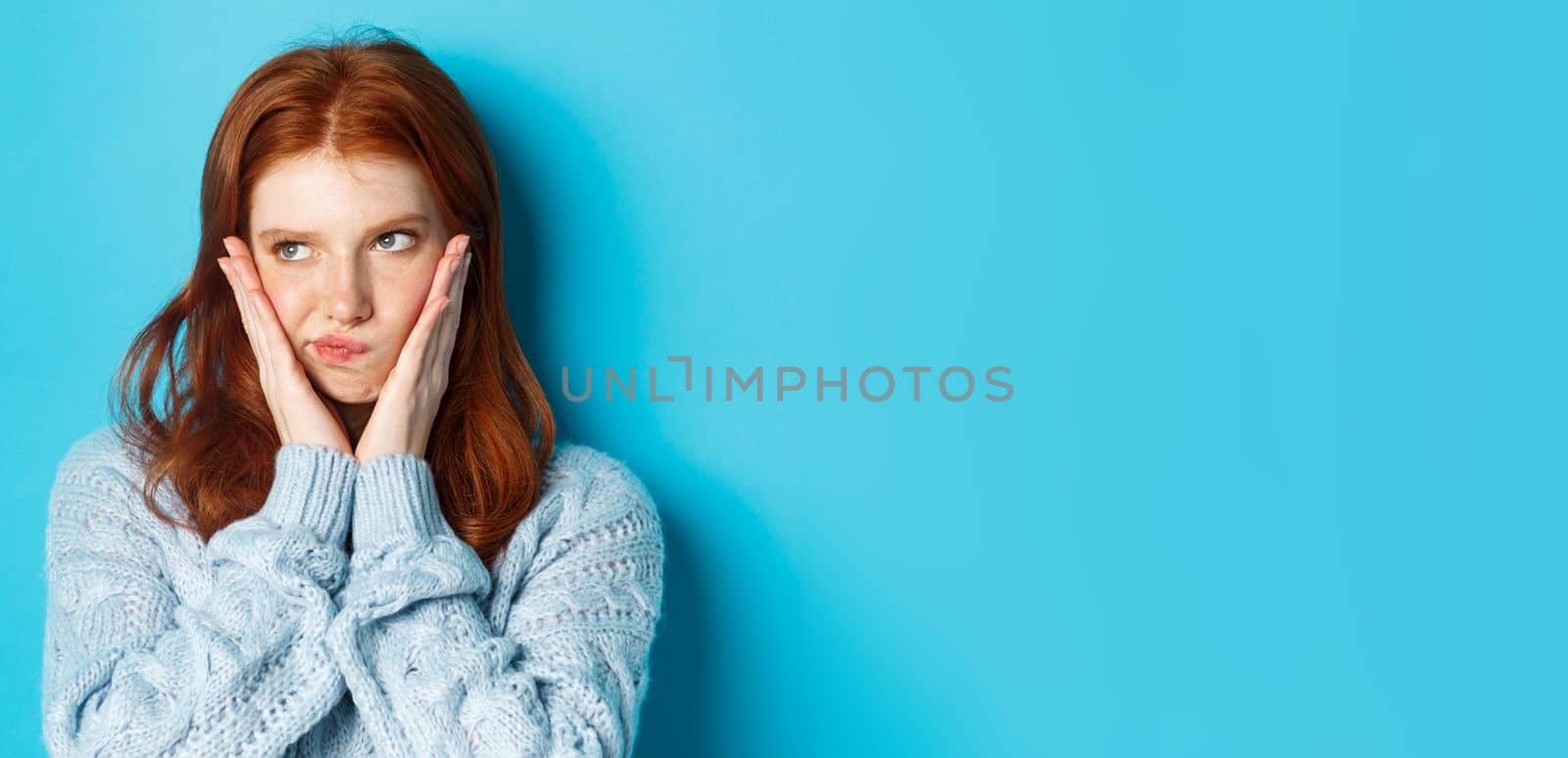 Close-up of troubled teenage girl with red hair staring left, pouting and looking grumpy at promo, standing over blue background.