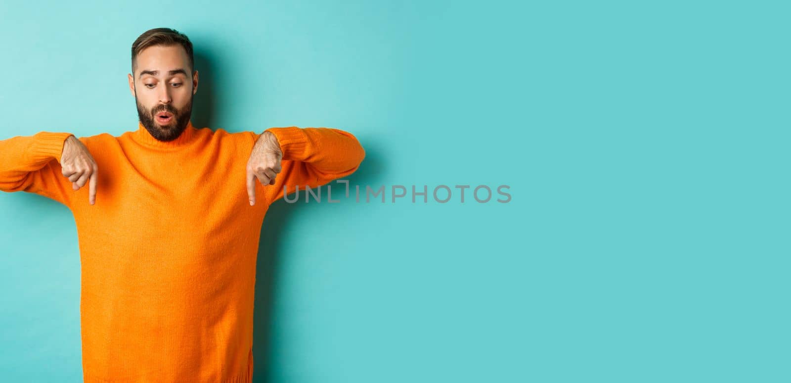 Amazed guy in orange sweater, pointing fingers down and looking with interest at promo, holiday discounts, standing over turquoise background by Benzoix