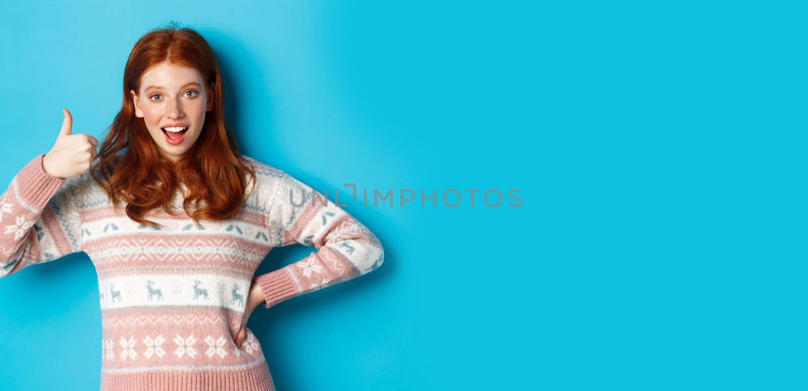 Excited redhead girl showing her support with thumb up, smiling pleased, saying yes in approval, agreeing, standing over blue background.