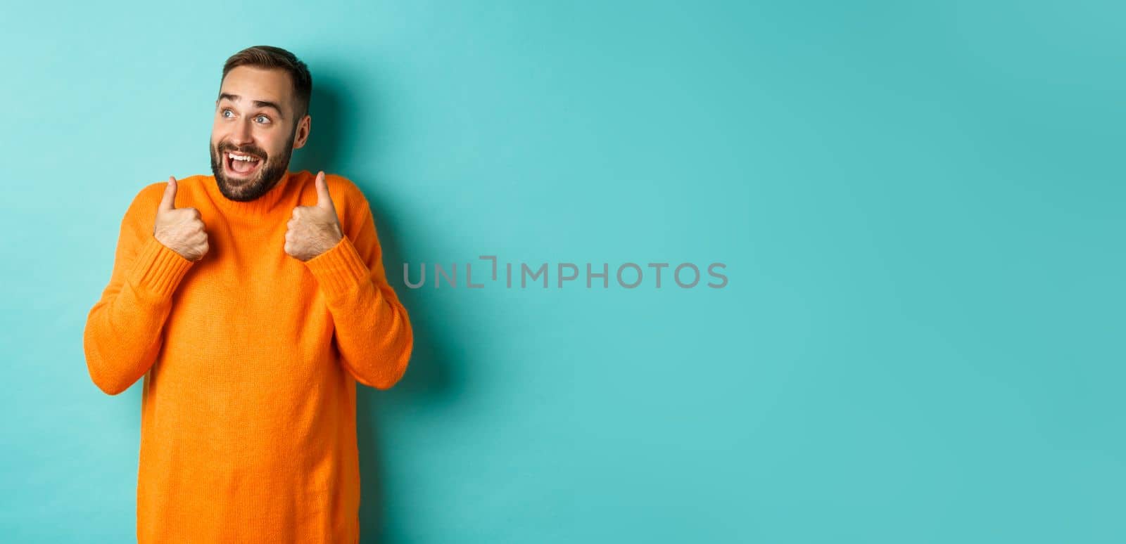 Cheerful man praising and looking at upper left corner, showing thumbs up amazed, super cool product, standing over light blue background.