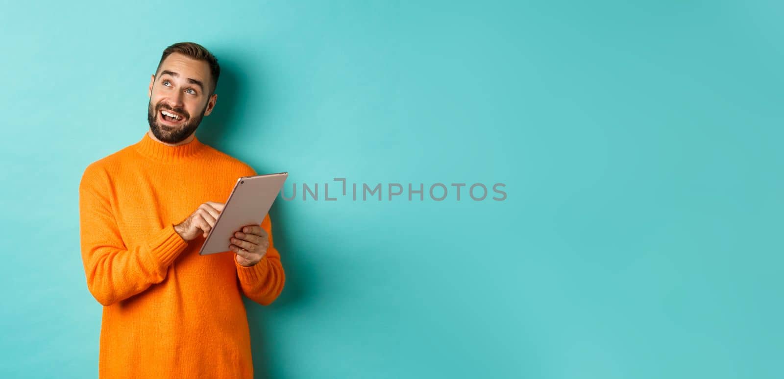 Handsome man thinking and using digital tablet, shopping online and pondering ideas, looking at upper left corner, standing over light blue background.