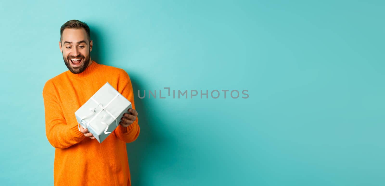 Holidays and celebration concept. Excited man receiving gift, looking happy at present and smiling, standing over blue background.