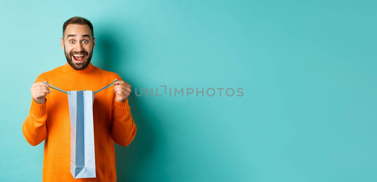 Surprised man open shopping bag and looking amazed, receiving gift on holiday, standing over turquoise background by Benzoix