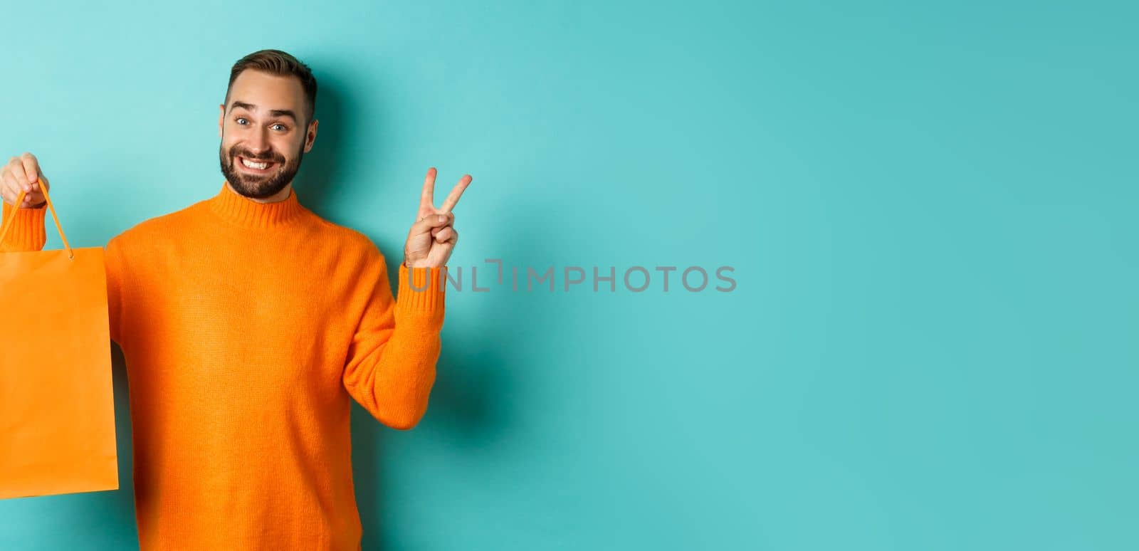 Happy young man showing peace sign and orange shopping bag, smiling pleased, standing over turquoise background. Copy space