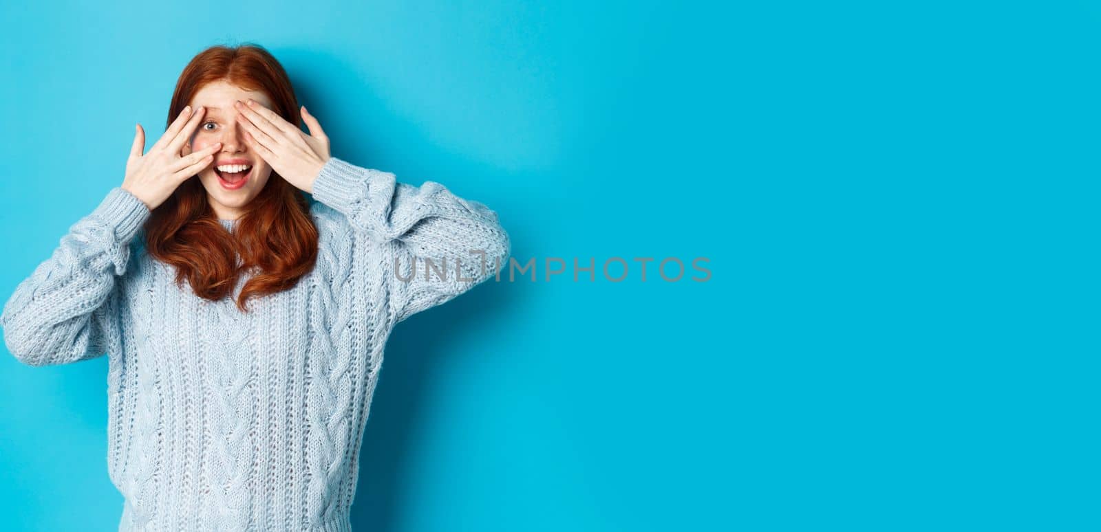 Excited teenage redhead girl open eyes to see holiday surprise, receiving presents, looking amazed at camera, standing over blue background by Benzoix