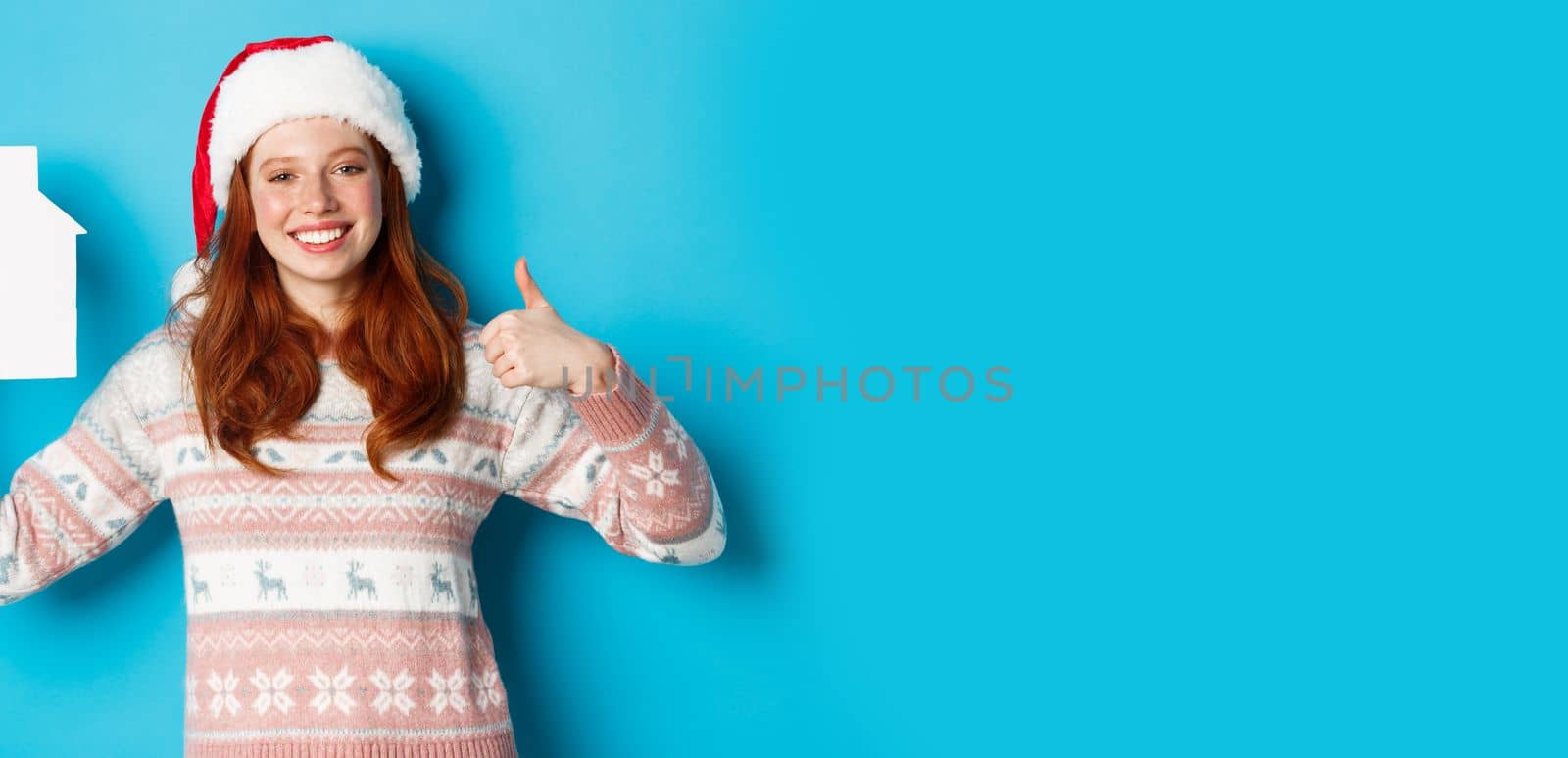 Holiday promos and real estate concept. Satisfied female model with red wavy hair, wearing santa hat and sweater, showing paper house model and thumbs-up, blue background.