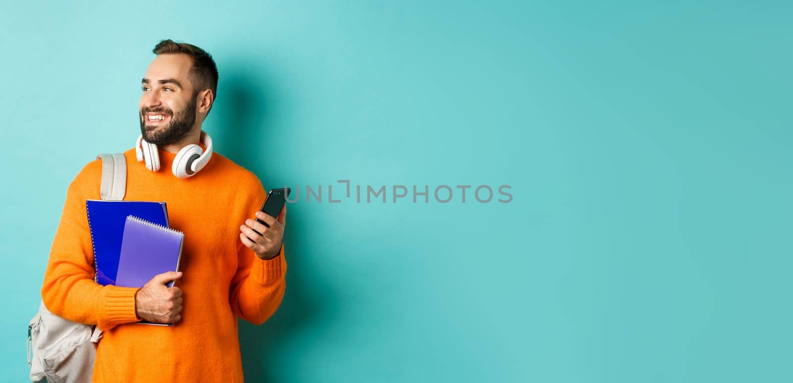 Education. Handsome male student with headphones and backpack, using mobile phone and holding notebooks, smiling happy, standing over turquoise background by Benzoix