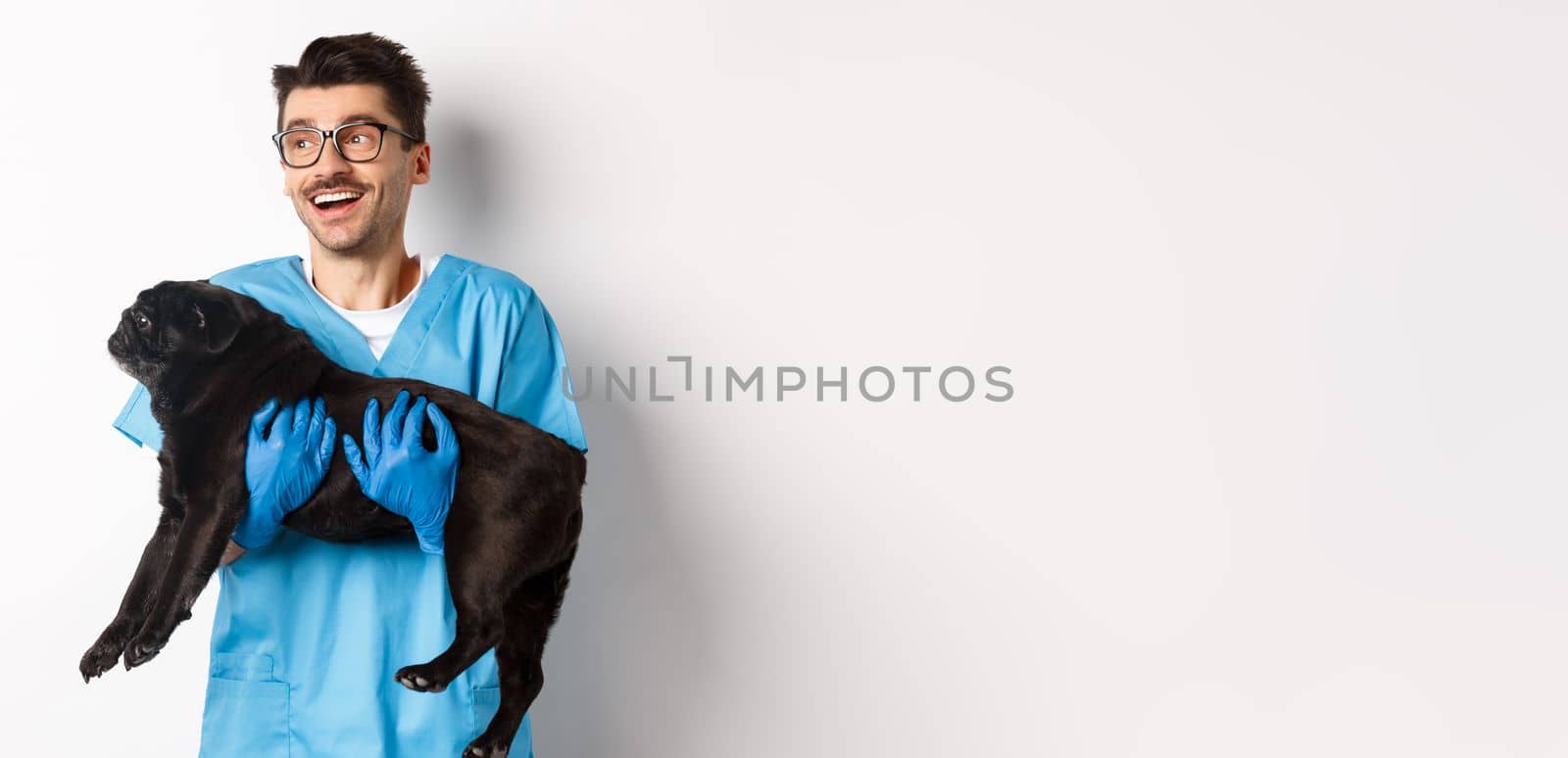 Vet clinic concept. Happy male doctor veterinarian holding cute black pug dog, smiling and looking left, standing over white background by Benzoix