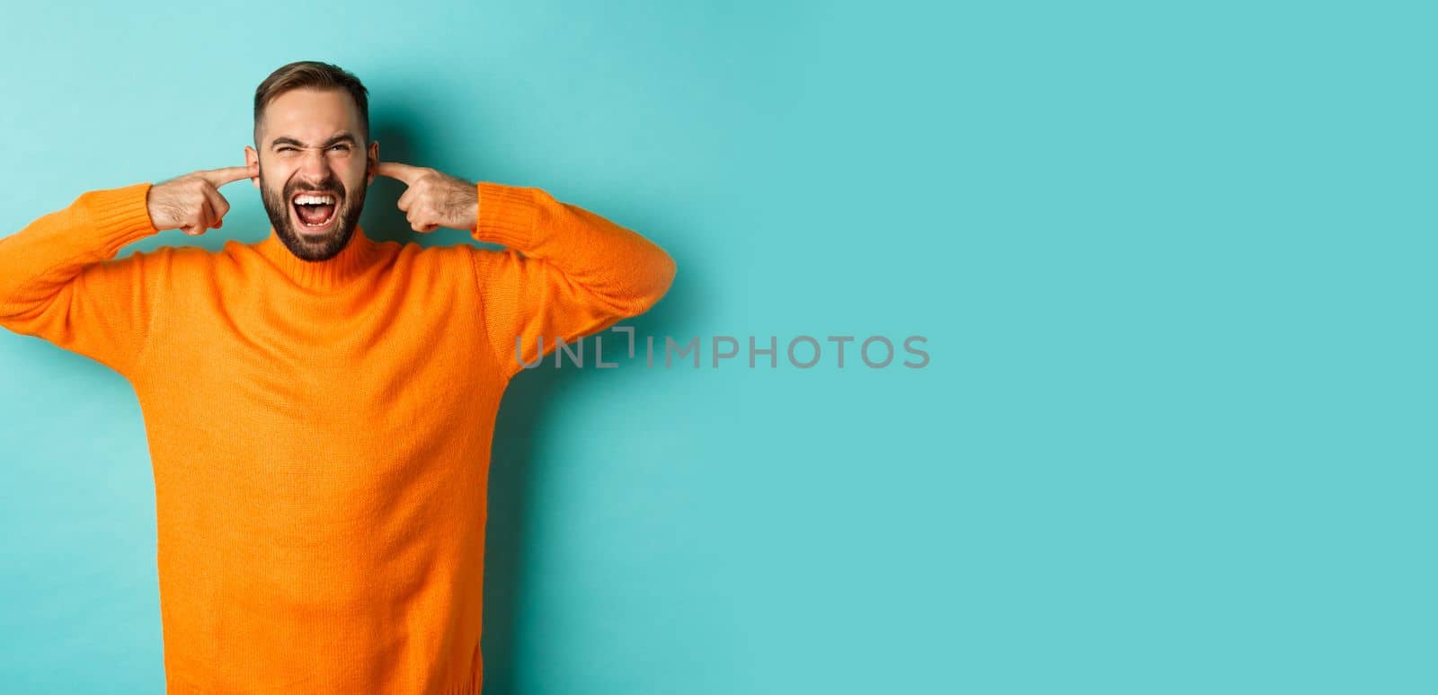 Displeased man shut ears and grimacing, loud neighbours, standing annoyed with noise, shouting angry and distressed, light blue background.