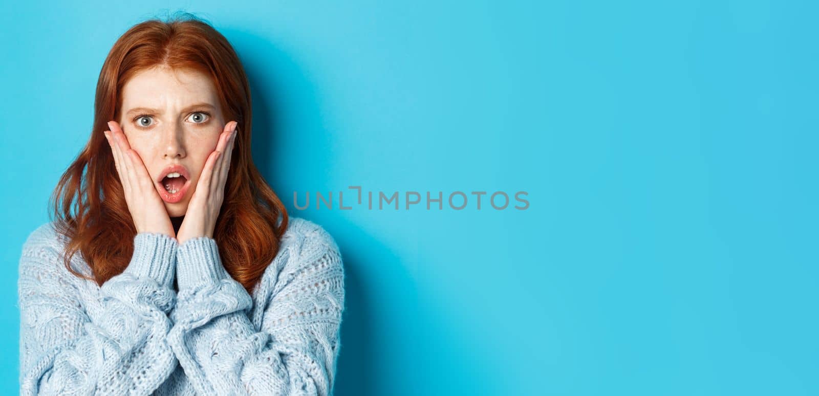 Close-up of shocked redhead girl staring at something displeasing, holding hands on face and gasping, standing over blue background.