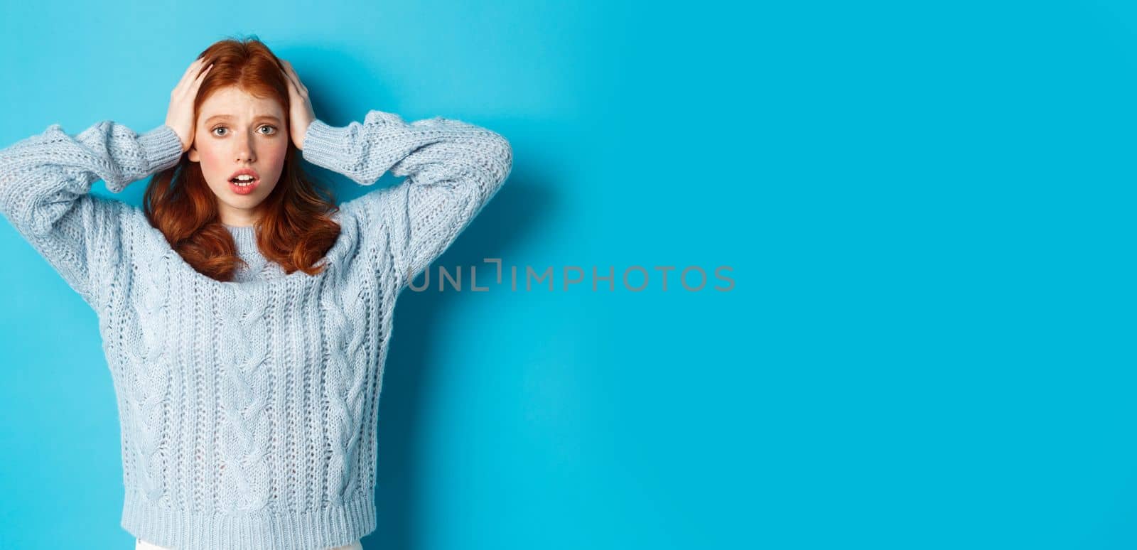 Nervous redhead girl standing overwhelmed, holding hands on head in panic and staring at camera, standing anxious against blue background.